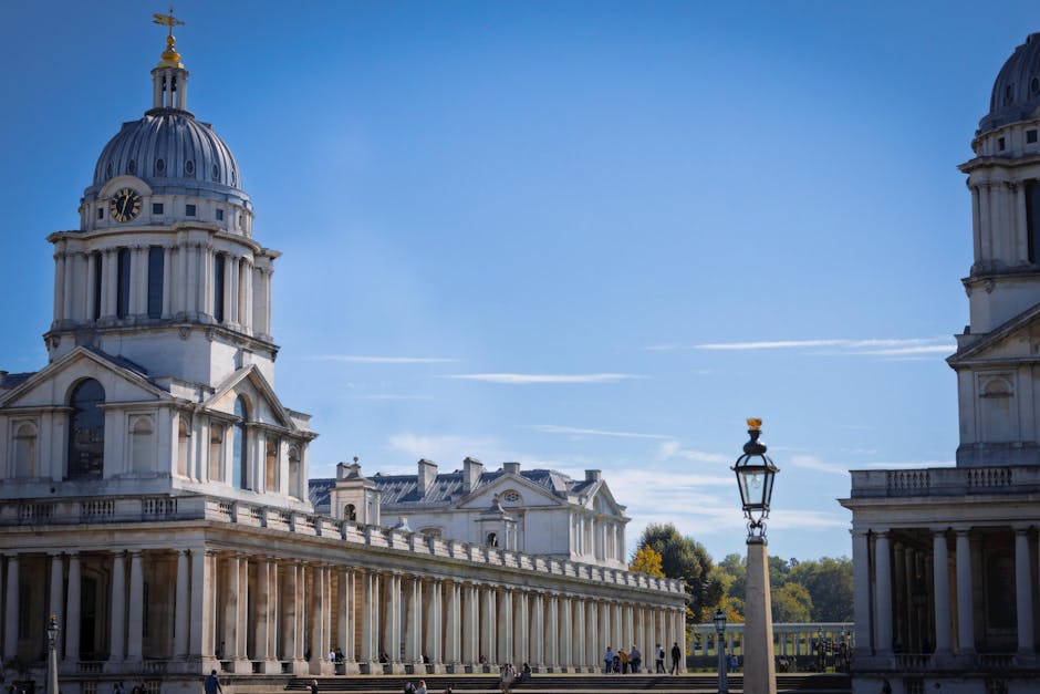 The image features a historic architectural structure in a European city, characterized by its large, ornate dome with a golden weathervane on top and a clock face visible on its façade. The building’s exterior is made of light-colored stone, displaying classical elements such as columns, decorative molding, and a series of evenly spaced arched windows. In the foreground, there is a vintage-style street lamp with intricate metalwork, positioned on a cobblestone or paved surface, while behind the building, a row of trees with green foliage can be seen. The sky above is clear and bright, with a few thin contrails streaking across the blue background. The scene suggests an area with pedestrian activity, possibly in a historical or cultural district, where private or independent waste collection services such as Waste Collection Greenwich could perform on-site clearance or rubbish removal, supported by the surrounding urban environment and the absence of visible waste containers or refuse. The overall composition emphasizes the building’s grandeur and the meticulous urban setting, aligning subtly with professional rubbish removal service contexts without explicit promotional language.
