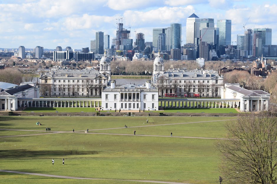 A wide view of a historic white neoclassical building with symmetrical architecture, including columns, domed towers, and decorative facades, situated within a large, well-maintained green lawn with multiple walking paths and trees in the foreground. The building is adjacent to a modern city skyline featuring tall glass skyscrapers and under-construction high-rise buildings in the background, with some construction cranes visible. The sky is partly cloudy with patches of blue, casting soft natural light over the scene. The image captures a contrast between the classical architecture of the historic estate and the contemporary urban development behind it, illustrating the surrounding environment often associated with private estate grounds or alternative waste handling sites, possibly serviced by independent rubbish collection services, with no visible waste or debris in the scene.