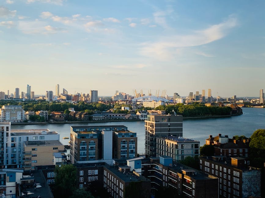 A wide view of a cityscape during daytime showing a river flowing through the urban area, with modern high-rise buildings in the foreground made of glass, concrete, and brick, some with balconies and flat rooftops, and mid-rise structures. In the background, a cluster of taller skyscrapers with various architectural styles, including one with a distinctive sloped roof and another with a curved glass facade, dominate the skyline. The river's surface reflects the sky, which is mostly clear with a few scattered clouds, and the surrounding environment is lush with green trees lining the riverbanks and interspersed among the buildings. The overall atmosphere is calm, with natural light illuminating the scene, highlighting the textures of the buildings and the gentle ripples on the water. This scene subtly relates to independent waste removal services, such as those provided by Waste Collection Greenwich, especially in urban settings where on-site clearance or private disposal may complement municipal rubbish collection options.