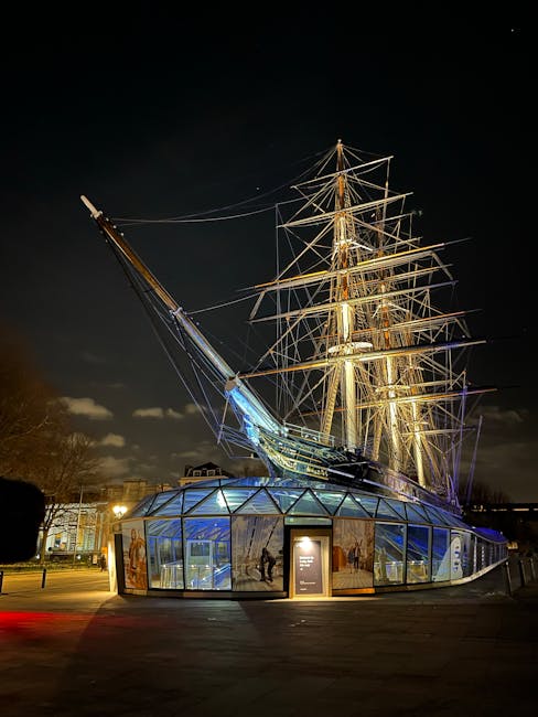 A historical maritime sculpture resembling a large sailing ship with detailed rigging and masts, illuminated at night with golden and blue lighting. The ship is mounted on a modern glass and metal structure that curves around its base, creating a futuristic appearance. The scene is set outdoors with a dark sky, and surrounding trees and buildings faintly visible in the background. The lighting highlights the intricate details of the ship's wooden textures and metal rigging, contrasting against the sleek, transparent glass enclosure at the bottom. While primarily a public art installation, it is located in an environment where it could be associated with displaying maritime history or culture, but it is not directly related to rubbish removal services. The overall scene emphasizes the contrast between historical design and contemporary lighting, captured in a clear night setting.