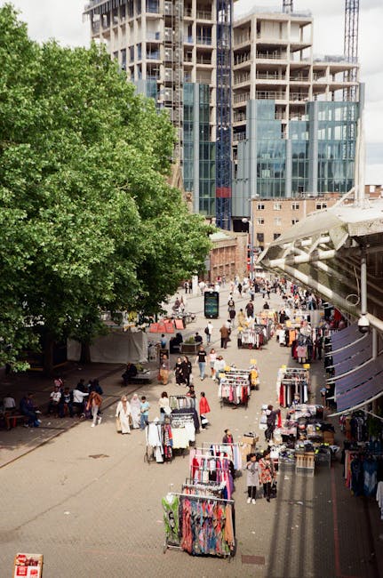 A busy outdoor market scene in an urban setting, with multiple clothing stalls arranged in rows on paved ground. Each stall is covered with tarps or fabric canopies, displaying a variety of garments such as dresses, shirts, and trousers hanging on racks. Several shoppers are browsing and walking between the stalls, some carrying shopping bags. To the left, a large leafy green tree provides partial shade over part of the market, contrasting with the surrounding modern high-rise buildings under construction in the background. The buildings have glass facades and visible concrete frameworks, indicating ongoing development in the area. On the right side, a structure with a white awning extends over part of the market, possibly a walkway or additional vendor space. The scene is well-lit by natural daylight, capturing the vibrant activity typical of an independent market, which may require regular rubbish collection and waste disposal to manage the assortment of clothing and other debris generated in such busy environments.