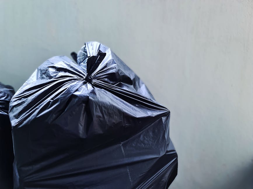 Close-up view of a large black plastic trash bag filled with waste, situated in the foreground against a plain, light-colored wall. The bag's crinkled, shiny surface reflects light, revealing creases and folds. The bag appears tightly sealed at the top with a gathered tie or knot, and it is positioned on the ground or a flat surface, suggesting it is ready for collection or disposal. In the background, partially visible, are additional black rubbish bags stacked or lined up, indicating a collection area for waste transfer or rubbish removal. The environment appears to be an indoor or sheltered space, possibly a designated waste storage area or behind a property, in a typical setting where private rubbish collection services like Waste Collection Greenwich could operate. The scene underscores an aspect of on-site waste management, reflecting the process of rubbish accumulation prior to scheduled collection, aligned with services in Greenwich for same-day or efficient waste disposal.