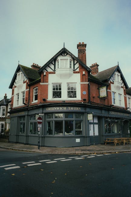 The image depicts a multi-story, traditional pub building situated at a street corner, with the signage reading 'Greenwich Tavern' on the dark-colored facade on the ground floor. The building features a combination of red brickwork and white-painted sections, with prominent timber framing details and a steeply pitched roof with multiple gables. Several large windows with white frames are visible on both the upper and lower levels, allowing natural light into the premises. The ground floor has a curved frontage with large glass panes, providing a clear view of the interior, while the exterior wall is painted in a muted dark tone. The surrounding environment includes a paved sidewalk, a street with dashed white lines, and a small pole with a traffic sign indicating a no-entry restriction for vehicles traveling in one direction. A wooden picnic table is positioned near the right side of the building, and a lamppost can be seen near the corner. The overall scene suggests an urban area where food and drink establishments like public houses are common, with Waste Collection Greenwich possibly providing rubbish removal services for such venues as part of their local waste management options. The sky is overcast with soft, diffuse lighting, emphasizing the building's details and materials without harsh shadows.