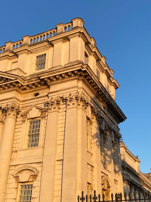 Close-up view of an ornate, classical-style building constructed from light-colored stone. The facade features decorative columns with detailed capitals supporting a prominent cornice with intricate dentil molding. Large rectangular windows with grid-patterned panes are visible on the upper floors, set within recessed frames. The structure has a highly detailed architectural design, with visible shadows cast by the sunlight, highlighting the textured stone surface. The building is situated outdoors under a clear blue sky, and part of a wrought-iron fence is visible at the lower right corner, indicating a private or historic property. The overall scene emphasizes the building's craftsmanship and classic European architectural elements, consistent with structures that might be associated with private estates or significant civic buildings, which could relate indirectly to the context of specialist waste management or on-site clearance for similar historic sites managed by waste collection services like Waste Collection Greenwich.