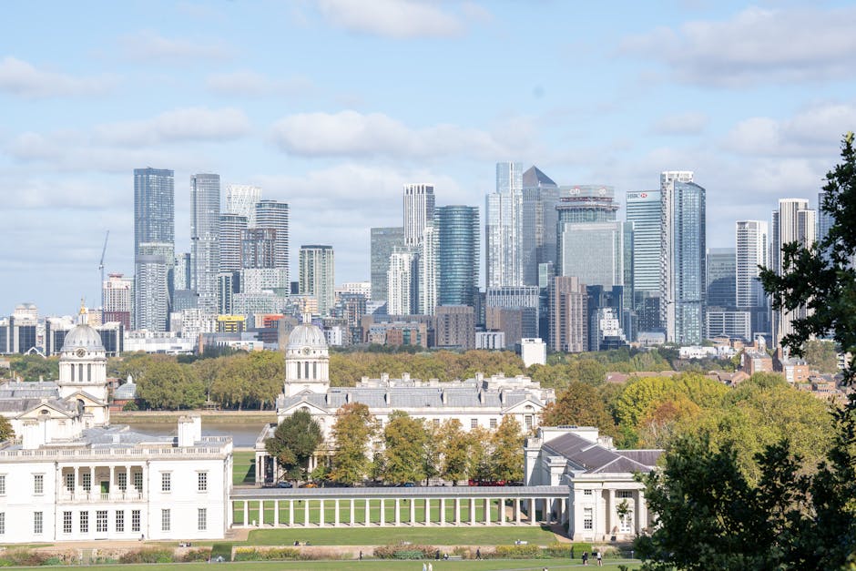 A panoramic view of a modern city skyline featuring numerous high-rise office buildings and residential towers made of glass and steel with reflective surfaces. In the foreground, there is a historic white building with classical architectural elements, including ornate columns and domed towers, situated within a park area with green lawns and scattered trees displaying early autumn foliage. The scene is set on a clear day with partly cloudy skies, casting soft shadows across the buildings. The environment suggests an urban setting with a blend of contemporary skyscrapers and traditional architecture, typical of city centres that might require private waste clearance or alternative rubbish removal solutions. Waste Collection Greenwich provides expert rubbish services that can facilitate efficient on-site clearance in areas with tight access, similar to the confined spaces near historic structures or densely built city zones, ensuring clean and accessible environments amid busy metropolitan landscapes.