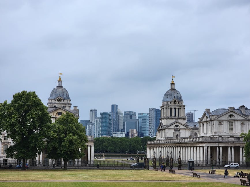 A wide view of an urban park shows two historic stone buildings with domed roofs and classical architectural details in the foreground. The building on the left features a grey stone facade, a large clock on its front, and a golden weather vane atop its dome. The building on the right has similar classical elements with a columned entrance and a decorative pediment. Between the buildings, a low iron fence encloses a grassy area with a few trees, including a prominent leafy green tree on the left side, partially obscuring the building. In the background, a cluster of modern skyscrapers with glass facades rises into a cloudy sky, contrasting with the historic structures. The scene includes a paved walkway with a few pedestrians, benches, and a parked vehicle near the fence, suggesting a city park setting. The subdued lighting and overcast weather create a neutral, professional atmosphere, consistent with an environment where private rubbish or waste management services, such as wastecollectiongreenwich.co.uk, might operate to handle waste collection delays or alternative disposal methods in urban areas.